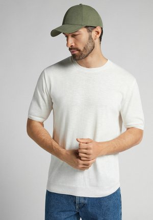 Pull blanc à manches courtes avec une finition texturée, porté avec un jean en denim bleu et une casquette de baseball vert olive. Pas de motifs ou d'accents notables.