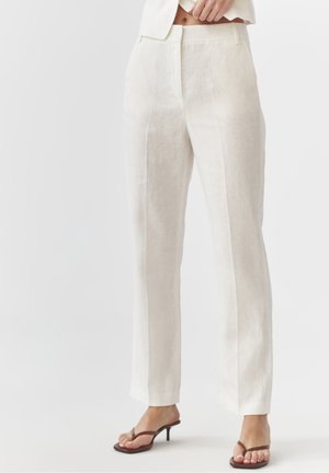 Mujer con pantalones beige de talle alto y pierna recta con pliegue frontal y sandalias marrones de tacón tipo tanga, de pie frente a un fondo blanco liso.