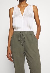 Haut blanc sans manches avec un col en V et des détails plissés, associé à un pantalon vert olive à cordon de serrage en tissu léger.