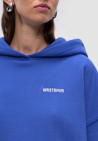 Felpa blu realizzata in tessuto morbido con un cappuccio prominente. Presenta la scritta ricamata in bianco "WRSTBHV" sul davanti. Design semplice, texture liscia.