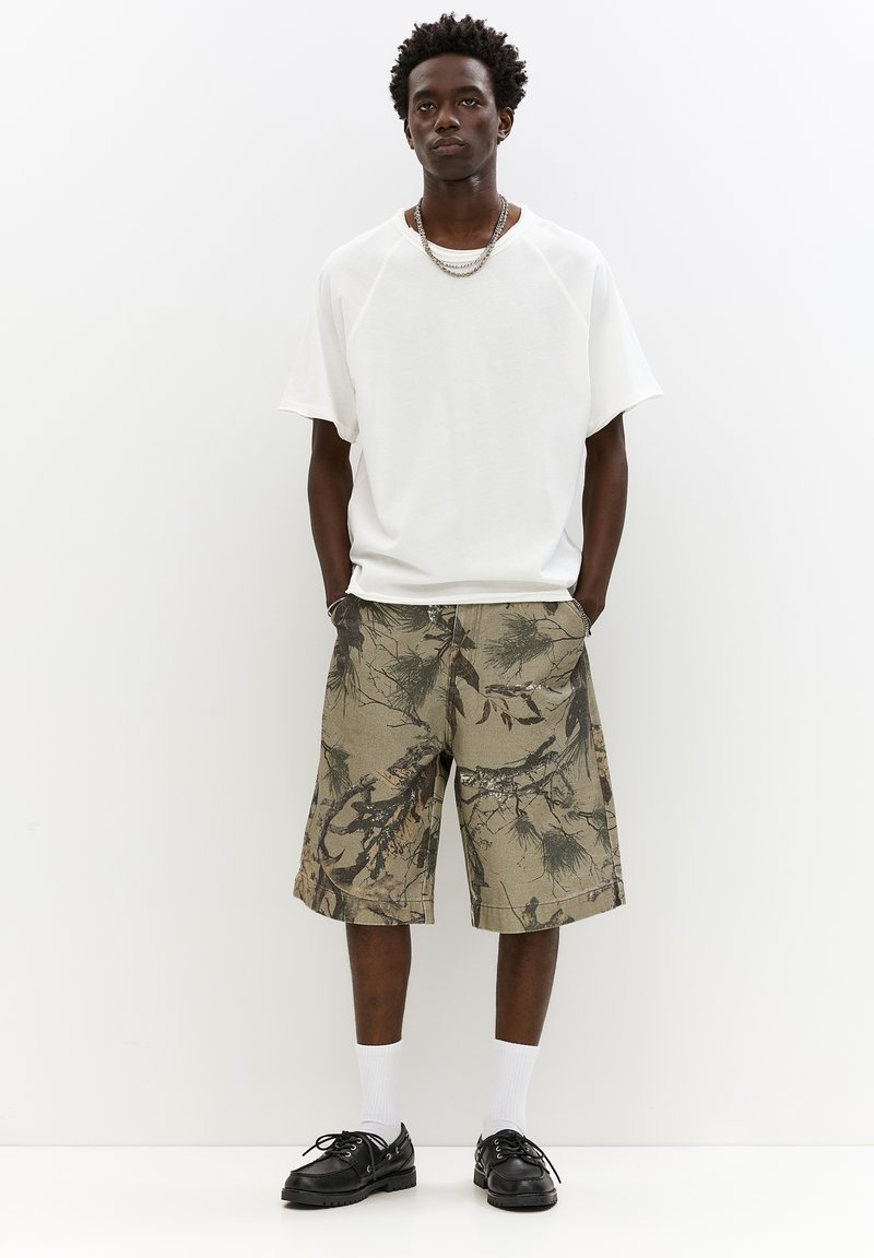 Hvid oversized t-shirt, khaki shorts med bladmønster, hvide sokker og sorte sko. Enkel design, afslappet pasform, velegnet til afslappet brug.