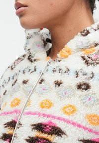 Felpa con zip in pile, caratterizzata da un morbido tessuto bianco testurizzato con motivi geometrici colorati in rosa, arancione e blu.