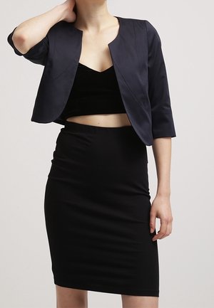 Femme portant un top court noir, un blazer court bleu marine avec des manches trois-quarts et une jupe crayon noire taille haute.