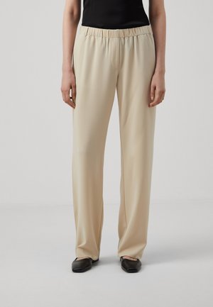 Beige wijde broek met elastische tailleband gedragen met een zwart topje en zwarte platte schoenen tegen een effen lichte achtergrond.