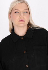 Camicia nera con texture, a maniche lunghe e colletto arrotondato, dotata di bottoni marroni e taschino sul petto. Il tessuto presenta un motivo a nido d'ape.