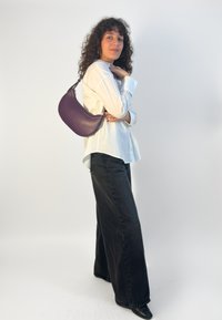 Fialová kožená kabelka na rameno s oblým tvarom, hladkou textúrou, striebornými kovaniami a nastaviteľným popruhom, nesená na bielej košeli a čiernych džínsoch.