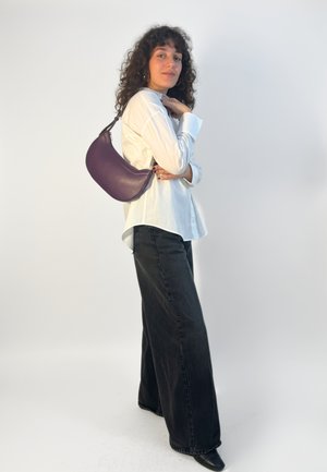 Sac à bandoulière en cuir violet avec une forme incurvée, une texture lisse, des accessoires en argent et une bandoulière réglable, porté avec une chemise blanche et un jean noir.