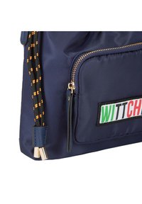 Zaino in tessuto blu navy con zip dorata, tracolla a motivo arancione e nero, caratterizzato da un'etichetta a patch con "WITTCH" in colori vivaci.