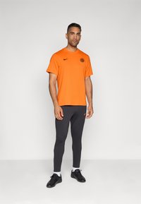 Camiseta naranja de Nike con logo, combinada con mallas gris ajustadas y zapatillas deportivas negras. Fondo simple.