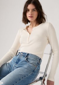 Top de manga larga en crema claro con textura de canalé y cuello henley, con botones metálicos, combinado con jeans de mezclilla azul de cintura alta.