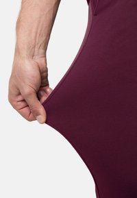 Bordeauxfarbener dehnbarer Stoff, der von einer Hand gezogen wird, zeigt eine glatte Textur und flexibles Material mit einer enganliegenden Passform.