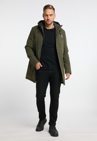 Olivgrön huva-jacka med grå foder, framdragkedja och tryckknappar; bärs över en svart skjorta och svarta jeans med svarta skor.