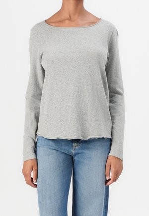 Personne portant un t-shirt à manches longues gris clair avec un col rond et un jean bleu en denim, debout devant un fond blanc.