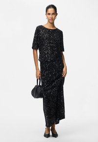 Mulher vestindo um top preto com lantejoulas e uma saia longa a condizer, segurando uma pequena bolsa preta, de pé contra um fundo branco.