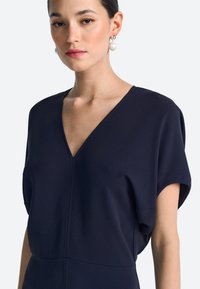 Granatowa sukienka z dekoltem w kształcie litery V i krótkimi rękawami. Miękka, teksturowana tkanina, z przeszyciami tworzącymi delikatne fałdy i smukłą sylwetkę.