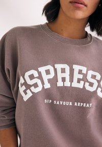 Felpa oversize in malva tenue con tessuto testurizzato. Presenta una scritta bianca bold "ESPRESS" e un testo più piccolo "SIP SAVOUR REPEAT" sotto.