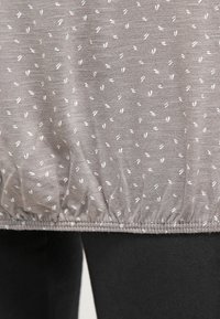 Světle šedý látkový top s bíle tečkovaným vzorem, který má na spodu nabraný lem. Měkká textura a lehký design.