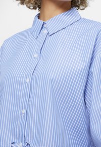Light blue striped shirt with a button-up collar, featuring white vertical stripes, smooth texture, and a tied detail at the hem.