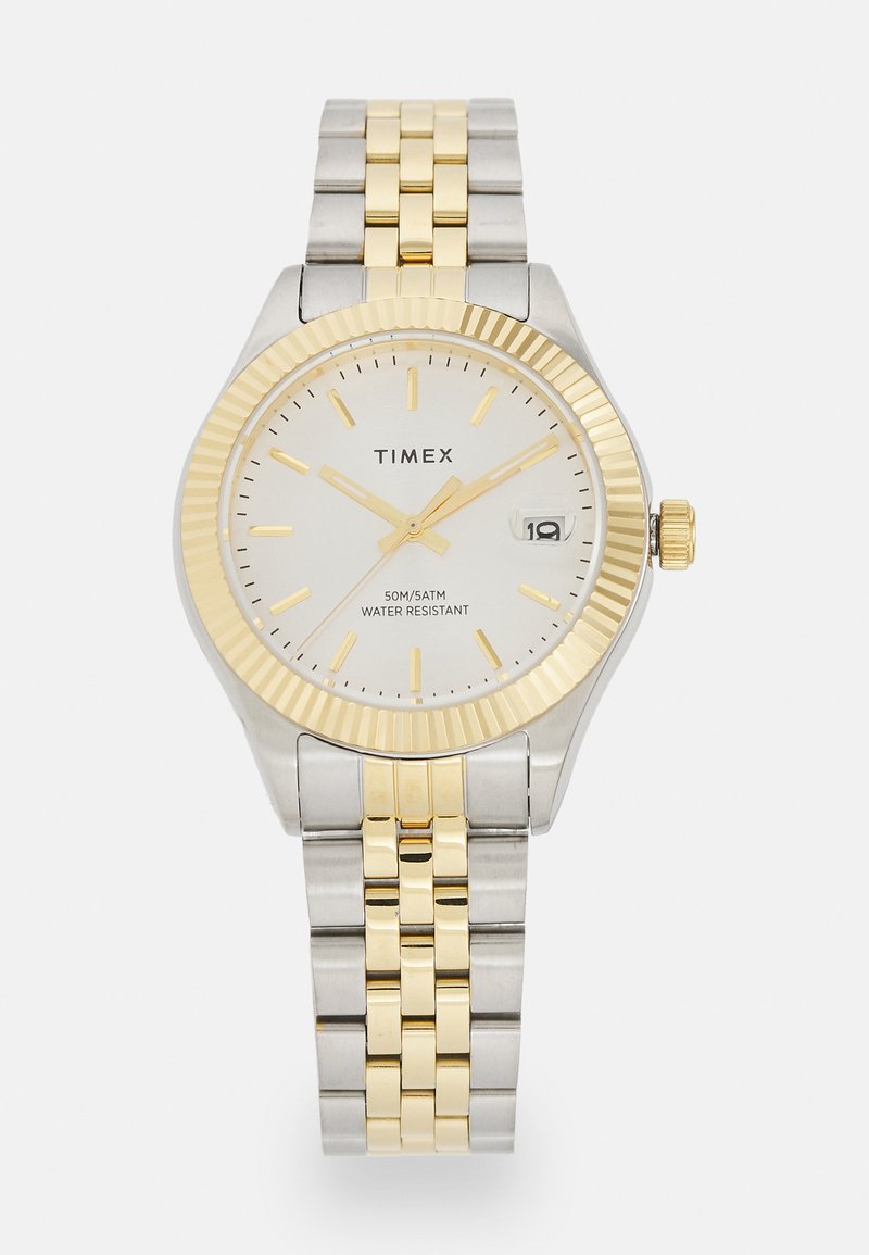 Montre Timex en argent et or avec une lunette texturée, un cadran blanc et un affichage de la date. Dispose d'un bracelet en métal bicolore et d'une résistance à l'eau jusqu'à 50 m.