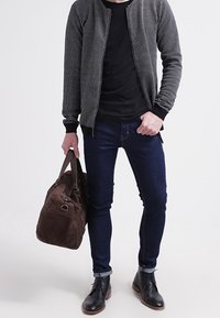 Svart bomberjacka med texturerat tyg, svart t-shirt, mörka jeans, brun mocka duffelväska och svarta läderstövlar.