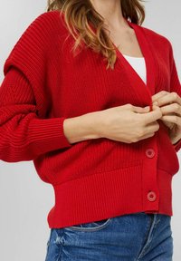 Roter gestrickter Cardigan mit V-Ausschnitt, überschnittenen Schultern und gerippten Bündchen und Saum. Er hat zwei Knöpfe in einer passenden Farbe.