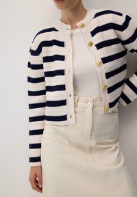 Mornarički i kremasti prugasti pletivi kardigan s zlatnim gumbima, okruglim izrezom i teksturiranim materijalom, uparen s svijetlom suknjom.