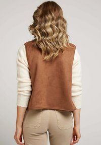 Femme avec des cheveux blonds ondulés portant un gilet sans manches marron par-dessus un pull crème et un pantalon beige, de dos contre un fond uni.