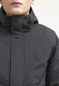 Il giubbotto a vento nero presenta un colletto alto, cordini regolabili e un logo impresso discreto. Il materiale appare liscio e leggero.