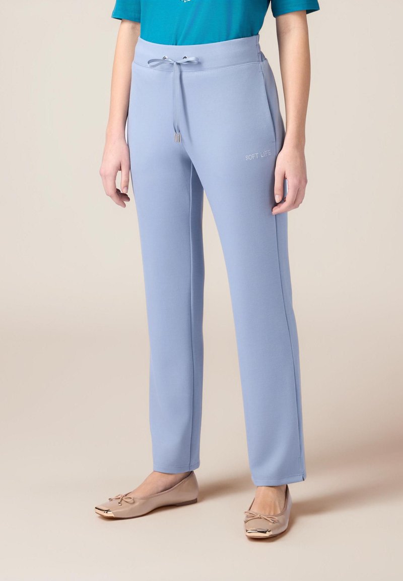 Pantalons de survêtement bleu clair avec une taille à cordon de serrage et du texte brodé "SOFT LIFE". Texture lisse, design à jambe droite, associés à des ballerines beiges.