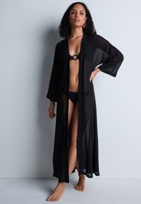 Kimono noir transparent avec de longues manches, ouvert à l'avant, et une texture légère. Porté par-dessus un bikini noir, mettant en valeur son design fluide.