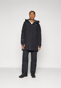 Osoba stoji u crnoj vodootpornoj jakni i hlačama, s osmijehom na neutralnoj pozadini, prikazujući odjeću pogodnu za vanjske aktivnosti.