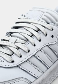 White leather sneakers with flat white laces, three parallel stripes on the side, and textured accents on the tongue and heel.