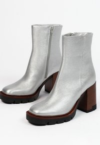 Zilveren metallic enkellaarzen met een gestructureerde afwerking, een zijrits en stevige bruine houten hakken. Rubberen zool met gripgroeven.