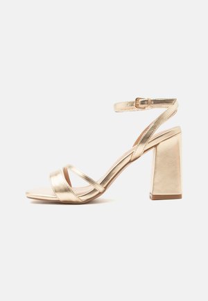 Guldmetallic højhælet sandal med blokhæl, to stropper foran og en ankelrem med spænde på hvid baggrund.