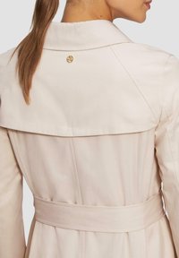 Lichtbeige trenchcoat, met een ceintuur in de taille, grote kraag en een decoratieve gouden knoop aan de achterkant. Glad, gestructureerd materiaal.