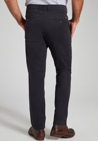 Pantaloni scuri navy realizzati in tessuto liscio, dotati di due tasche posteriori con bottoni e gamba dritta su misura, abbinati a scarpe in pelle marroni.