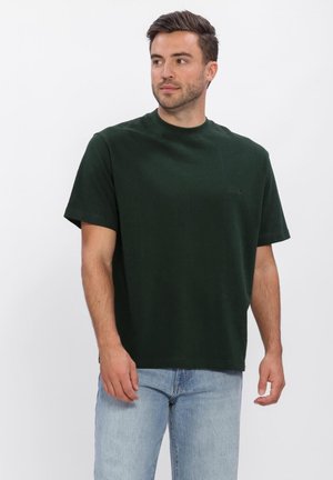 Man met kort donker haar, die een donker groen T-shirt met korte mouwen en lichtblauwe jeans draagt, staat tegen een effen witte achtergrond.