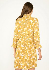 Vestido floral amarillo con cuello con volantes, mangas largas con puños acampanados y una cintura ajustada con una falda fruncida. Fondo blanco.