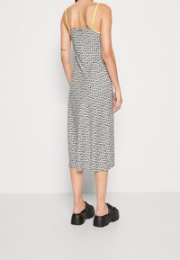 Femme portant une robe midi à fleurs noires et blanches avec des bretelles jaunes, debout, vue de dos, chaussée de chaussures plateformes noires sans lacets.