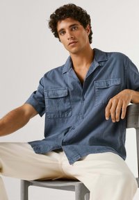 Joven con cabello rizado, vistiendo una camisa azul de manga corta abotonada y pantalones claros, sentado en una silla contra un fondo liso.