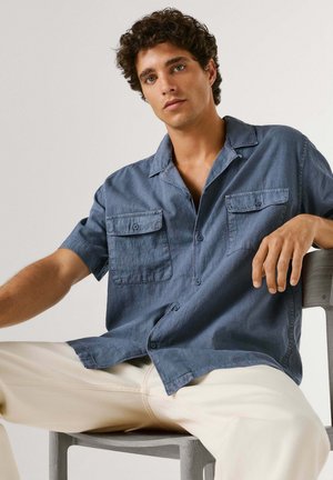 Młody mężczyzna z kręconymi włosami, ubrany w niebieską, krótką koszulę na guziki i jasne spodnie, siedzi na krześle na tle jednolitego tła.