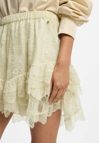 Mujer que lleva una falda de encaje color crema con capas con volantes y una cintura elástica, combinada con una blusa de satén beige de manga larga.