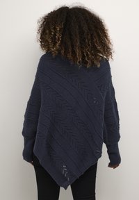 Mörkblå stickad poncho med diagonala texturmönster, lös passform och vida ärmar. Har en längre bakre fåll och subtila openwork-detaljer.
