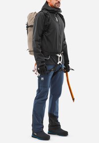 Veste softshell noire, pantalon bleu avec poches zippées, gants, harnais d'escalade, piolet orange et sac à dos beige ; les détails incluent un tissu texturé et un design ergonomique.