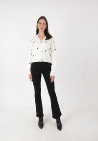 Witte cardigan met zwarte hartpatronen, knoopsluiting aan de voorkant en geribde manchetten. Gecombineerd met zwarte flared jeans en scherpe zwarte laarzen.