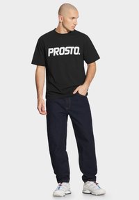 Czarna koszulka z krótkim rękawem z białym logo "PROSTO.". Połączona z ciemnoniebieskimi dżinsami i białymi butami sportowymi. Prosty, casualowy design.