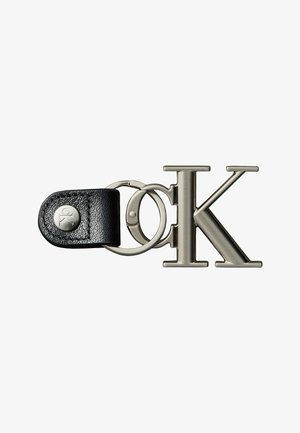 Metalen sleutelhanger met zwarte leren band en zilveren knop, met grote gepolijste letters "C" en "K" verbonden door een metalen ring.