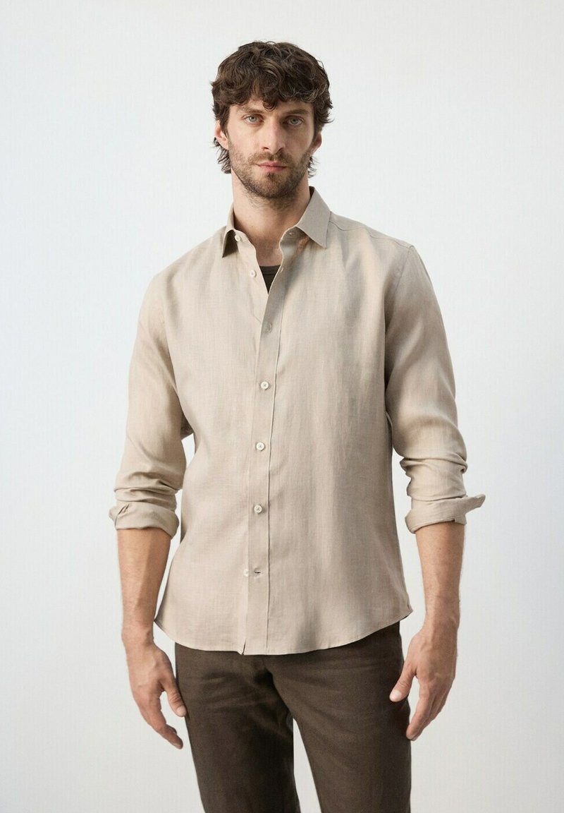 Homme aux cheveux bouclés bruns portant une chemise beige à manches longues avec les manches retroussées et un pantalon marron foncé, debout devant un fond uni.