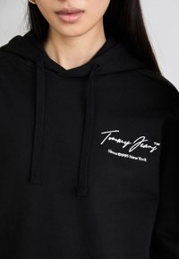 Felpa nera con cappuccio e coulisse, caratterizzata da un logo bianco con la scritta "Tommy Jeans" sul petto. Tessuto morbido e vestibilità rilassata.