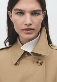 Trench coat beige con design doppiopetto, ampio colletto e bottoni marroni, indossato su una camicia bianca con colletto alto. Tessuto di consistenza liscia.
